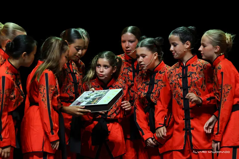 Des filles en vestes kung-fu rouges brodées avec micros serre-tête entourent un livre illustré tenu par l'une d'elles