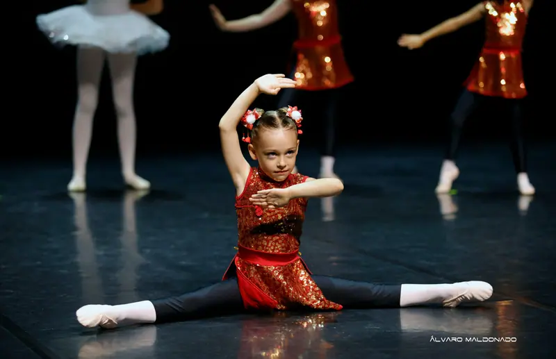 Une fillette en robe rouge à paillettes et leggings noirs fait le grand écart au sol, un bras arrondi au-dessus de la tête