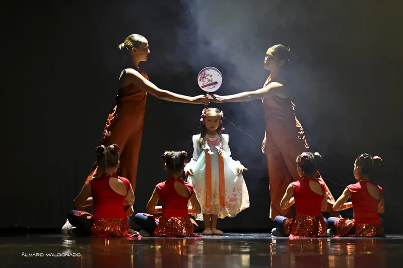 Deux danseuses en combinaisons rouille tiennent un éventail rond peint au-dessus d'une fillette en robe pastel, entourée de filles en rouge