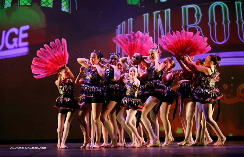 Des filles en costumes de chat léopard posent avec de grands éventails de plumes roses devant un décor de cabaret aux néons