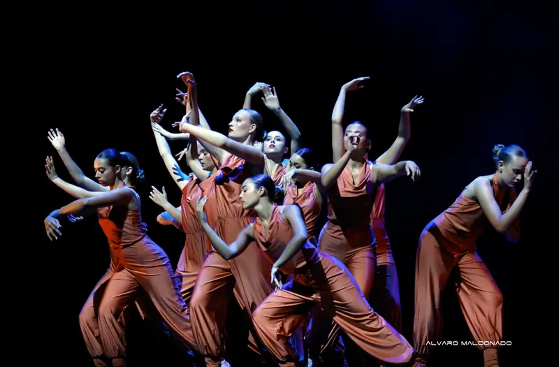 Danseuses adolescentes en combinaisons satinées rouille bougent ensemble bras levés sous un projecteur sur une scène sombre