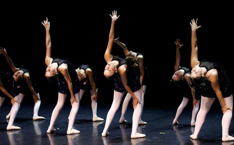 A line of girls in dark sequinned leotards and pale tights arch backwards with one arm raised on a dark stage